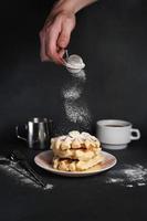 kaukasisch des Mannes Hand Sträusel Zucker Pulver auf köstlich Waffeln, Karamell Soße, Kaffee Tasse, Milch, Dessert Löffel, Sieb auf schwarz Beton Hintergrund foto