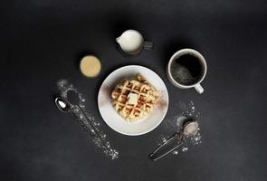 oben Aussicht von lecker Waffeln Platte, Karamell Soße, Kaffee Tasse, Milch, Dessertlöffel, Sieb auf ein schwarz Beton Hintergrund foto