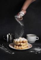 Frau bereit Frühstück, köstlich Waffeln, Karamell Soße, Kaffee Tasse, Milch, Dessert Löffel, Sieb auf schwarz Beton Hintergrund foto