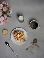 oben Aussicht von lecker Waffeln Platte, Karamell Soße, Kaffee Tasse, Milch, Dessertlöffel, Sieb, Rosa Blumen auf ein grau Beton Hintergrund foto