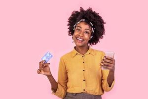 Porträt von ein afrikanisch Frau zeigen Anerkennung Karte und Handy, Mobiltelefon Telefon. jung Frau Einkaufen online. schön Frau lächelnd suchen beim Kamera foto