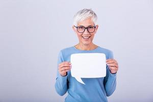 schön reifen Frau mit Brille halten Rede Blase mit ihr Hände und lächelnd beim das Kamera , isoliert Über grau Hintergrund foto