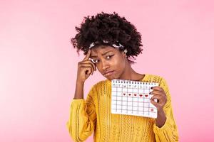 jung afrikanisch amerikanisch Frau halten Menstruation Kalender Über isoliert Hintergrund mit Überraschung Gesicht. Frau halten Menstruation Kalender mit verwirrt Gesicht, Angst und aufgeregt mit Angst Ausdruck foto