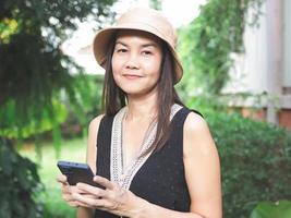 asiatisch Frau, tragen Hut und schwarz oben ärmellos, Stehen im das Garten halten Handy, Mobiltelefon Telefon und suchen beim Kamera. foto