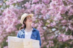 asiatisch Frau Tourist halten Stadt Karte während Gehen im das Park beim Kirsche blühen Baum während Frühling Sakura Blume Festival Konzept foto