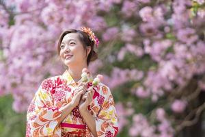 japanisch Frau im traditionell Kimono Kleid halten Süss Hanami Dango Dessert während Gehen im das Park beim Kirsche blühen Baum während Frühling Sakura Festival mit Kopieren Raum foto