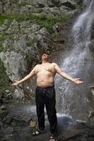 ein Alten Mann steht beim ein Berg Wasserfall, Verbreitung seine Waffen und genießt das Moment. foto