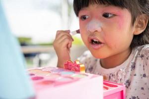 bezaubernd wenig Kind asiatisch Mädchen Farben ihr Mund mit Rosa Kinder Köpfe und sieht aus im das Spiegel. ein Kind Theaterstücke beim Zuhause im ein Spielzeug Schönheit Salon. erhöhen, ansteigen Lernen Entwicklung zum Vorschulkinder. foto