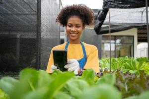 Porträt von glücklich halb thailändisch Hälfte afrikanisch Frau Farmer Stehen hinter ein Gemüse Handlung und mit Smartphone zum prüfen Befehl. Konzept von Landwirtschaft organisch zum Gesundheit, vegan Essen und klein Geschäft. foto