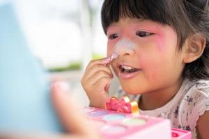 bezaubernd wenig Kind asiatisch Mädchen Farben ihr Mund mit Rosa Kinder Köpfe und sieht aus im das Spiegel. ein Kind Theaterstücke beim Zuhause im ein Spielzeug Schönheit Salon. erhöhen, ansteigen Lernen Entwicklung zum Vorschulkinder. foto