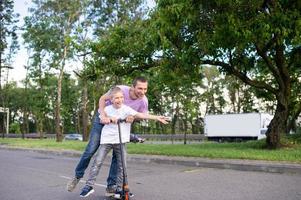 ein Vater Fahrten seine Sohn im ein Weiß T-Shirt auf ein Roller, das glücklich Kind Verbreitung seine Hände foto