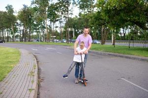 ein Vater Fahrten seine Sohn im ein Weiß T-Shirt auf ein Roller, das glücklich Kind Verbreitung seine Hände foto