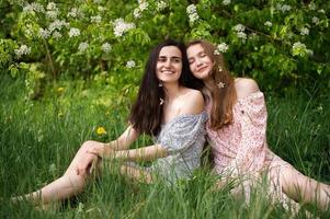 zwei jung Mädchen im Kleider sind Sitzung auf das Grün Gras unter ein Weiß Baum und Lachen foto