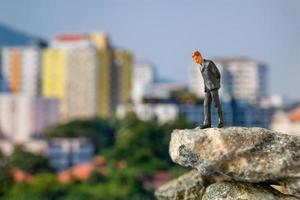 Miniaturgeschäftsmann, der auf einem Felsen mit Gebäuden im Hintergrund steht foto