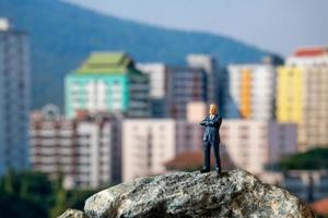 Miniaturgeschäftsmann, der auf einem Felsen mit Gebäuden im Hintergrund steht foto