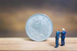 Miniaturgeschäftsleute, die nahe einer Münze mit einem hölzernen Hintergrund, Geschäftskonzept stehen foto