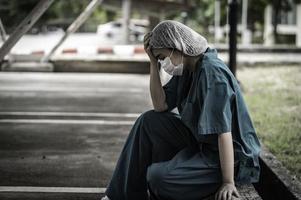 müde depressive asiatische krankenschwester trägt gesichtsmaske blaue uniform sitzt auf dem krankenhausboden, junge ärztin gestresst von harter arbeit foto