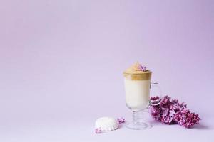schön Dalalgona trinken ein schaumig Kaffee im ein transparent Becher und Blumen lila zu lila il lila Hintergrund. Nächster zu das Becher ist ein Mäusespeck und ein Ast von lila. Kopieren Raum