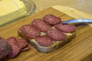 Butter, ein Scheibe von Brot mit Butter, geschnitten Salami und Messer auf ein Schneiden Tafel. foto