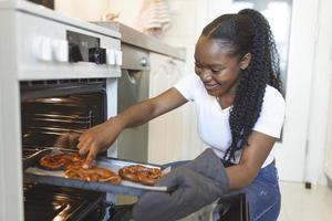 jung afrikanisch amerikanisch Frau im Küche Backen frisch Brezel, inländisch Leben foto