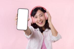 glücklich heiter lächelnd asiatisch Frau mit kabellos Kopfhörer zeigen leer Bildschirm Handy, Mobiltelefon Telefon oder Neu Smartphone Musik- Anwendung Werbung Attrappe, Lehrmodell, Simulation isoliert auf Rosa Studio Hintergrund. foto