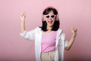 glücklich jung asiatisch Frau Modell- mit stilvoll modisch Sonne Brille genießen Hören Musik- durch Kopfhörer Audio- und Tanzen isoliert auf Rosa Studio Hintergrund. Technologie, Mädchen Mode, Zubehörteil Konzept. foto