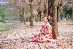 jung Dame im traditionell Kimono Kleid Praktiken Methoden Ausübungen Meditation beim Kirsche blühen Baum. Emotion Lächeln foto