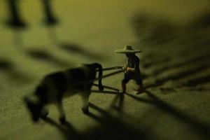 Miniatur Zahl von ein Farmer Pflügen ein Feld mit seine Kuh auf ein Karton. foto