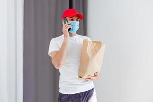 Lieferung Mann halten Papier Tasche mit Essen auf Weiß Hintergrund, Essen Lieferung Mann im schützend Maske foto