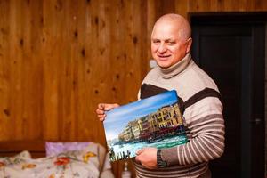 Alten Mann halten ein Foto Segeltuch im ein hölzern Haus