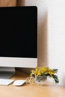 Home Office mit Computer, ein Strauß Mimosen in einer Vase. foto
