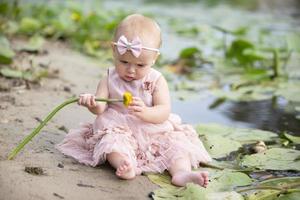 ein schön wenig Mädchen sitzt auf das Bank von das Fluss und hält ein Gelb Lilie im ihr Hände. Kind und Natur. foto