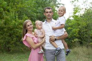 Mann und Ehefrau und ihr wenig Kinder. Familie Porträt im Natur. Mama und Papa sind halten ein Bruder und Schwester im ihr Waffen. ein jung Familie mit Kinder ist suchen beim das Kamera. foto