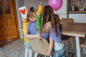 Töchter gratulieren ihr Mama auf Mutter Tag, ein Karte mit ein Herz, Blumen und ein Ballon beim Zuhause im das Küche, Sie alle Umarmung. Kinder Überraschung ihr Mutter zum das Urlaub. foto