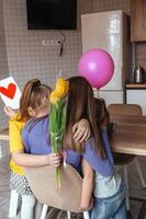 Töchter gratulieren ihr Mama auf Mutter Tag, ein Karte mit ein Herz, Blumen und ein Ballon beim Zuhause im das Küche, Sie alle Umarmung. Kinder Überraschung ihr Mutter zum das Urlaub.