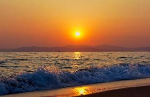 Wellen, die auf einem Strand mit orange bewölktem Sonnenuntergang über Bergen krachen