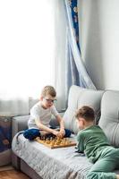 Jungs spielen hölzern Schach Lügen auf ein grau Sofa im Vorderseite von das Fenster foto