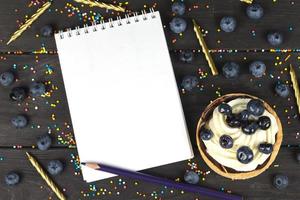 auf ein hölzern Tafel Lügen ein Kuchen mit Sahne und Blaubeeren und ein Weiß Notizbuch mit leer Seiten. foto