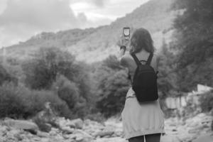 ein jung Tourist Frau mit schön wellig Haar, mit ein Rucksack auf ihr Schultern, Reisen durch das Berge in der Nähe von das Fluss und Fotografien Natur auf ihr Telefon, schwarz und Weiß Foto