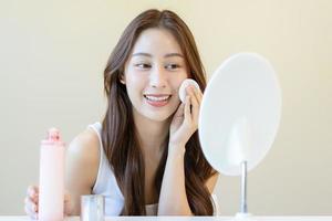 glücklich Schönheit, schön asiatisch jung Frau, Mädchen suchen im zu Spiegel, halten Baumwolle Pad, bewirbt sich Gesichts- wischen auf ihr Gesicht, entfernen bilden Vor Dusche im Badezimmer, Haut Pflege auf Weiß Hintergrund. foto