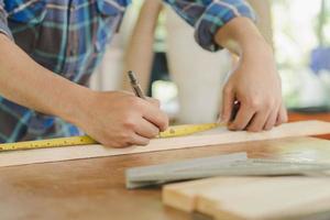 Zimmerei Fachmann, Hand von asiatisch jung Mann, männlich Zimmermann verwenden Band messen, Werkzeug auf Tafel Holz im klein Geschäft. Holzarbeiten, Möbel Herstellung, Konstruktion Industrie. Inhaber klein Geschäft Personen. foto