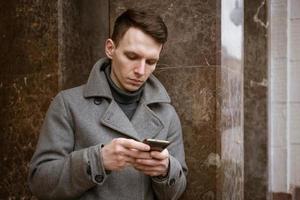 Nahansicht Schuss modisch jung Mann mit seine Handy, Mobiltelefon Telefon, tragen des Mannes Mantel foto