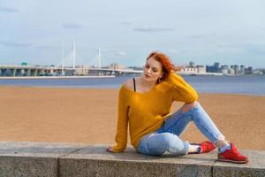 attraktiv jung Rothaarige Frau Sitzung draußen auf am Wasser, tragen Gelb foto