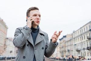 emotional jung Mann mit Handy, Mobiltelefon Telefon, tragen ein elegant grau Mantel, steht foto