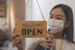 wieder öffnen, asiatisch jung Inhaber Einzelhandel, Kaffee Geschäft Frau drehen Zeichen Tafel öffnen tragen Gesicht Maske, schützen zu Pandemie von Coronavirus, wieder öffnen Geschäft nach schließen Sperrung Quarantäne im COVID-19, Neu normal. foto