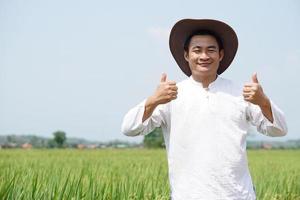 gut aussehend asiatisch Mann Farmer trägt Hut, Weiß Shirt, erziehen zwei Hände Daumen hoch, steht beim Paddy Feld. Konzept, Landwirtschaft Beruf. zufrieden im Pflanzen und landwirtschaftlich Produkt. thailändisch Bauer. foto