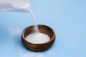 Zucker ist gegossen von ein Weiß Papier Tasche in ein hölzern Schüssel auf ein Blau Hintergrund foto