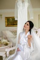 das Morgen von das Braut. Porträt von ein Brünette Braut im ein Weiß Seide Mantel hinter ein Kleid foto