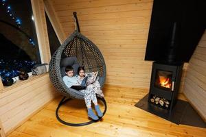 Bruder mit Schwester lesen Bücher im komfortabel hängend Stuhl gegen Kamin im gemütlich hölzern winzig Kabine Haus. Leben im Landschaft. foto