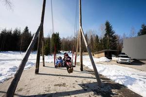 Mutter schwingen mit Töchter im groß hölzern schwingen im früh Frühling schneebedeckt Berge gegen winzig Haus und Wagen. foto